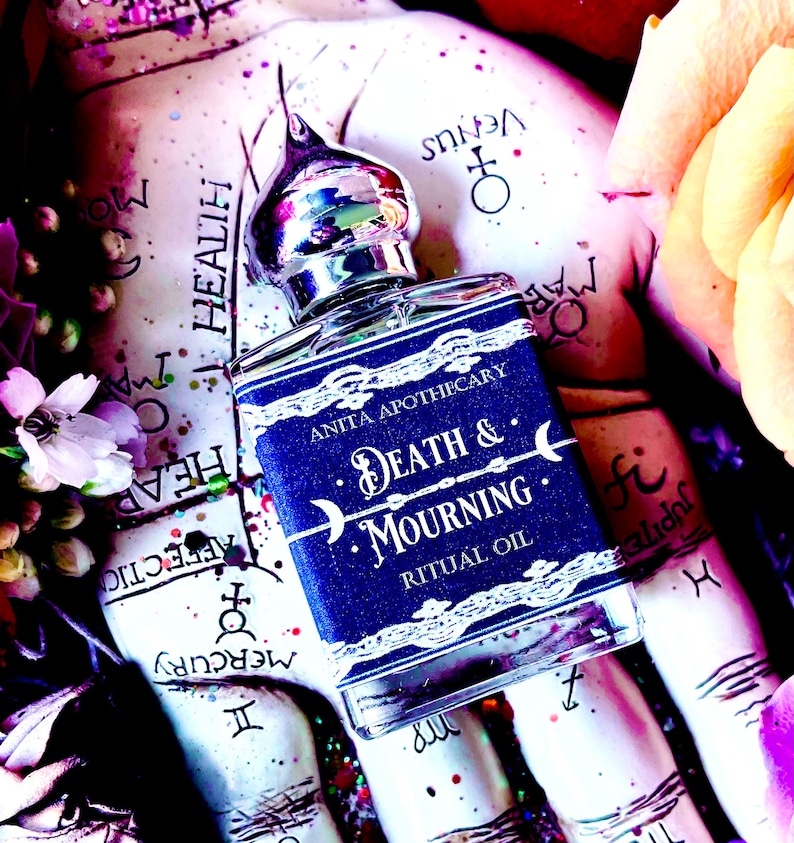 Pu&ograve; includere: Una bottiglia di vetro blu con un tappo argentato e un'etichetta bianca che recita "Anita Apothecary Death & Mourning Ritual Oil". La bottiglia &egrave; appoggiata su una superficie con uno sfondo colorato.