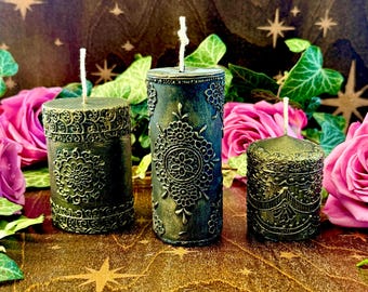 May include: Three green candles with intricate gold designs. The candles are on a brown wooden surface with a star pattern.