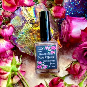May include: A clear bottle of crystal-infused nail polish with a black cap. The label reads "Mani Magick" and "Anita Apothecary" with floral accents. The background features pink roses and iridescent crystals. The bottle contains 15ml of polish.