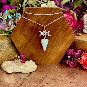 May include: A silver necklace with a pentagram charm and a moon phase pendant. The pendant is made of a white, opaque stone. The necklace is displayed on a hexagonal wooden stand.