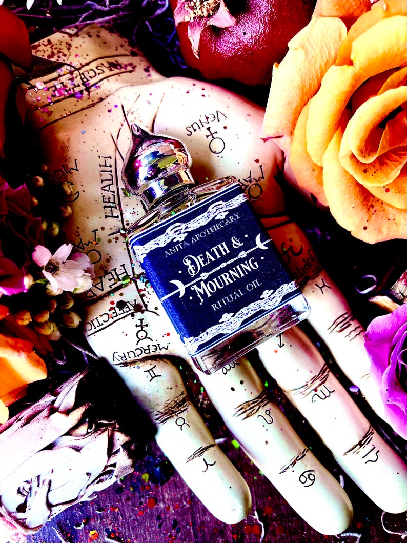 Pu&ograve; includere: Un piccolo flacone di vetro blu scuro con un tappo argentato e un'etichetta che recita "Anita Apothecary Death & Mourning Ritual Oil". Il flacone &egrave; tenuto in una mano con uno sfondo bianco che presenta simboli e segni astrologici.
