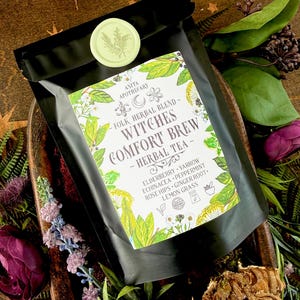 May include: A black bag of herbal tea labeled "Witches Comfort Brew" with a light green seal. The label includes the text "Anita Apothecary" and lists ingredients like elderberry and ginger root. The bag is surrounded by decorative flowers and foliage.