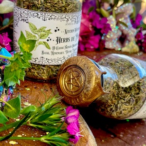 May include: Two glass jars filled with dried herbs, labeled "Kitchen Witch... Herbs de Provence... A Classic French Blend... Basil, Rosemary, Thyme". The jars are sitting on a wooden surface with pink flowers and glitter.