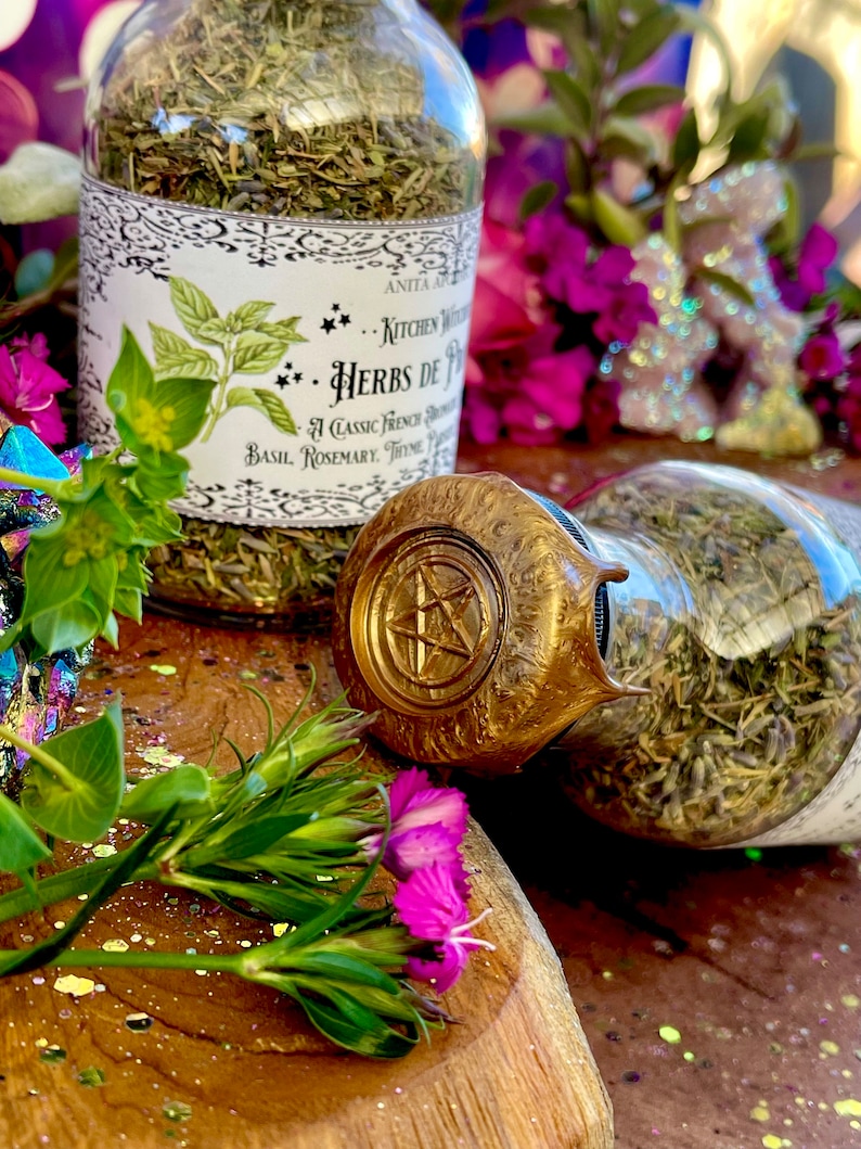 May include: Two glass jars filled with dried herbs, labeled "Kitchen Witch... Herbs de Provence... A Classic French Blend... Basil, Rosemary, Thyme". The jars are sitting on a wooden surface with pink flowers and glitter.