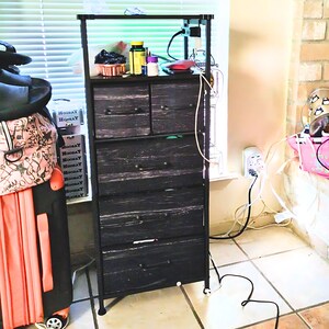 May include: A black metal 5-drawer dresser with a wood-grain finish. The dresser has a top shelf and is sitting next to a window. There are several cords and wires plugged into an outlet on the wall.