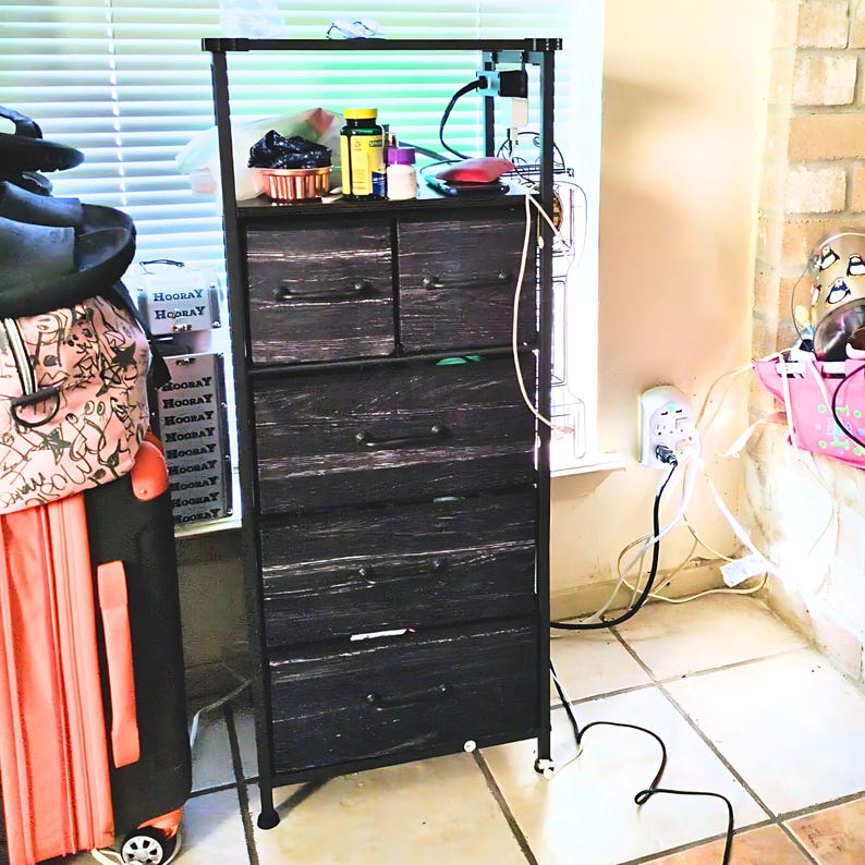 May include: A black metal 5-drawer dresser with a wood-grain finish. The dresser has a top shelf and is sitting next to a window. There are several cords and wires plugged into an outlet on the wall.