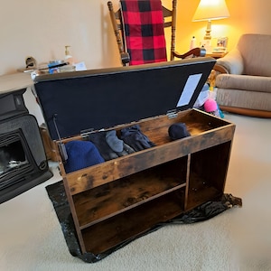 May include: A brown wooden storage bench with a black leather top and a lift-up lid. The lid is open and reveals a shelf with gloves and a hat inside. The bench has two shelves below the lid.