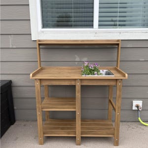 May include: A wooden potting bench with a stainless steel sink and two lower shelves. The bench has a top shelf and is filled with purple flowers. The bench is made of light brown wood.