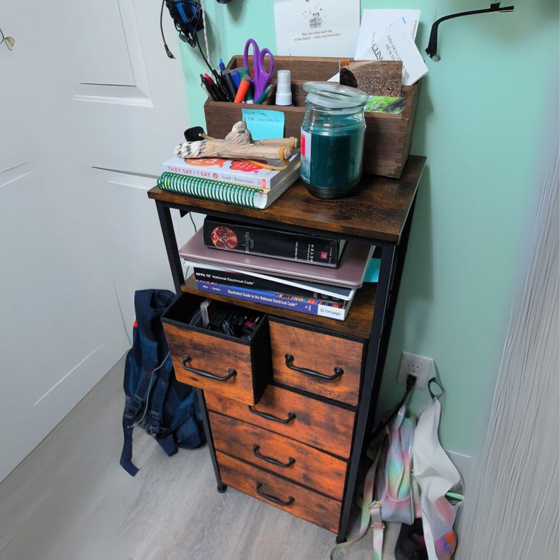May include: A black metal and wood 5-drawer dresser with a top shelf. The dresser has a rustic brown finish and is filled with books, a candle, and other items.