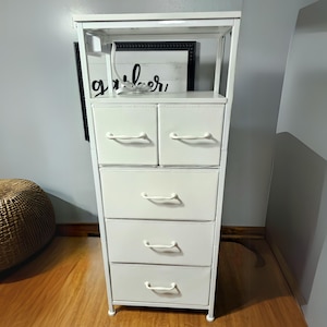 May include: White metal 5-drawer dresser with a shelf on top. The drawers have white handles and the dresser is standing on a wooden floor.