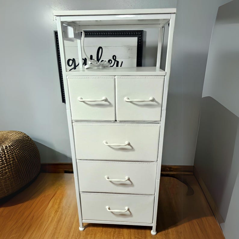 May include: White metal 5-drawer dresser with a shelf on top. The drawers have white handles and the dresser is standing on a wooden floor.