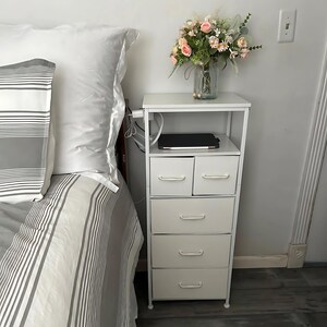 May include: White metal nightstand with five drawers, a top shelf, and a laptop on the shelf. The nightstand is next to a bed with a white and gray striped comforter.