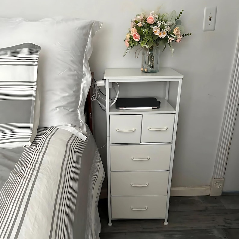 May include: White metal nightstand with five drawers, a top shelf, and a laptop on the shelf. The nightstand is next to a bed with a white and gray striped comforter.