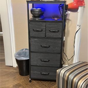 May include: A black metal and wood 5-drawer dresser with a top shelf. The dresser has a blue light underneath the top shelf. The dresser is on a wood floor.