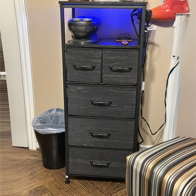 May include: A black metal and wood 5-drawer dresser with a top shelf. The dresser has a blue light underneath the top shelf. The dresser is on a wood floor.