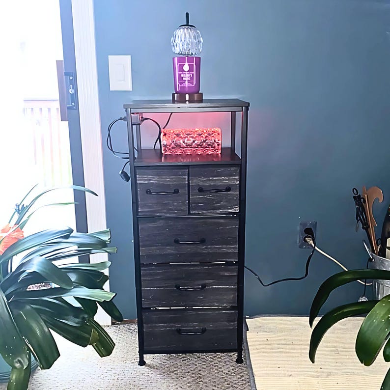 May include: A black metal 5-drawer dresser with a wood-grain finish. The top drawer has a clear glass lamp with a white globe and a purple candle on top. The dresser is lit with a red light underneath the top drawer.