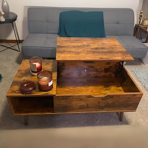 May include: A wooden coffee table with a lift-top and a drawer. The table has a distressed brown finish and is sitting on a gray rug. There are two candles and a bowl on the table.