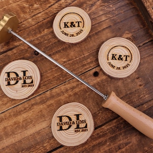 Puede incluir: Un hierro de marcar con mango de madera y cabeza de latón, junto a cuatro posavasos de madera. Los posavasos están grabados con iniciales, nombres y fechas. La veta de la madera es visible en los posavasos.