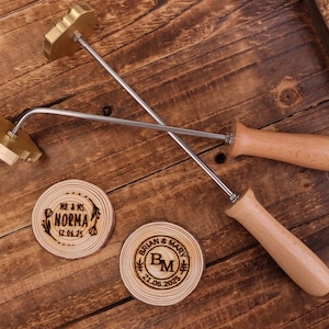 May include: Two wood-handled branding irons with brass heads are crossed on a wooden surface. Two engraved wooden coasters display names and dates. The coasters are light brown with dark brown text.
