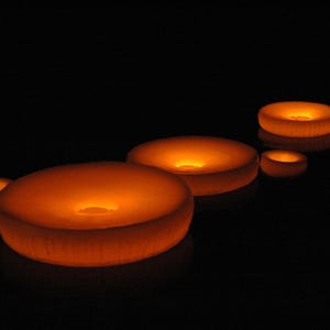 Puede incluir: Cinco objetos circulares de color naranja brillante de distintos tamaños dispuestos en un grupo sobre un fondo oscuro. Cada objeto presenta una superficie superior lisa y redondeada con una hendidura central, emitiendo luz cálida.