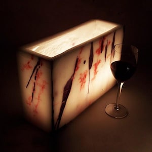 May include: A rectangular, translucent white stone ice bucket featuring irregular dark red and black streaks throughout its surface. A clear glass stemware filled with dark red liquid stands next to the illuminated, hollow stone container.