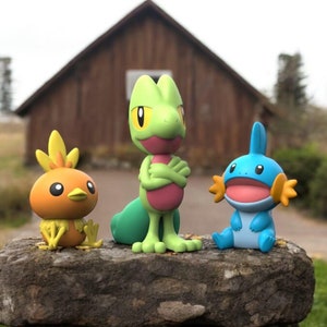 Peut inclure: Trois figurines Pokémon colorées : un petit oiseau orange avec des détails jaunes, un gecko vert avec les bras croisés et une créature bleue avec des nageoires orange. Elles sont posées sur un rocher, avec un bâtiment en bois flou en arrière-plan.