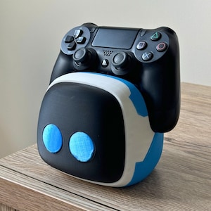 Op de afbeelding: Een zwart-witte 3D-geprinte controllerstandaard met blauwe accenten. De standaard is gevormd als een robotkop met twee blauwe ogen. Een zwarte PlayStation 4-controller rust op de standaard.