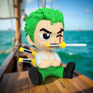 Peut inclure: Figurine au crochet de Zoro de One Piece, avec des cheveux verts, une chemise blanche et un pantalon vert. La figurine a une épée dans la bouche et deux épées derrière le dos. Elle est assise sur une surface en bois avec un arrière-plan flou de l'océan.