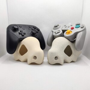 Op de afbeelding: Twee gamecontrollers, één zwart en één grijs, rusten op bijpassende schedelvormige decoratieve objecten. De controllers hebben verschillende knoppen en richtingspads. De schedels zijn gebroken wit en hebben een uitgesneden ontwerp.