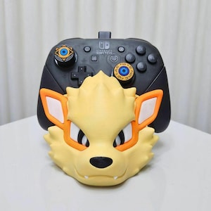 Op de afbeelding: Een 3D-geprinte Nintendo Switch-controllerstandaard met een geel en oranje Pokémon-personage met een zwarte neus en ogen. De controllerstandaard is zwart met een blauwe en gouden knop bovenop.