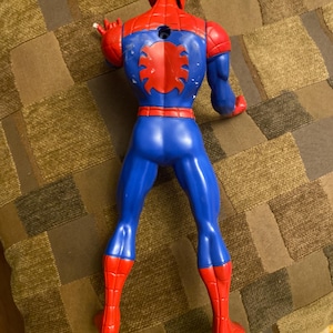 May include: A plastic Spider-Man action figure, viewed from the back. The figure is primarily red and blue, with a red spider emblem on the back. The figure's head and hands are red, and the legs are blue with red boots.