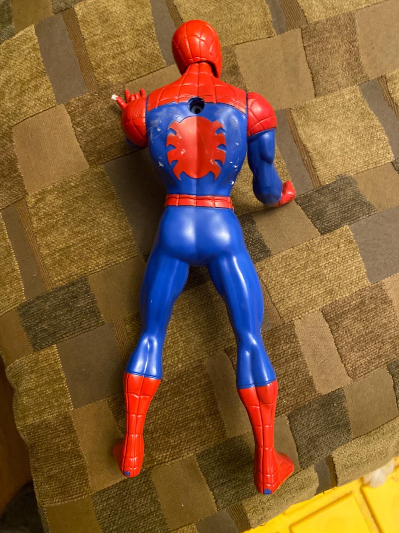 May include: A plastic Spider-Man action figure, viewed from the back. The figure is primarily red and blue, with a red spider emblem on the back. The figure's head and hands are red, and the legs are blue with red boots.