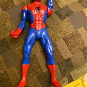 May include: A plastic Spider-Man action figure in a dynamic pose. The figure features a red and blue color scheme, with a red mask, torso, and boots, and blue arms and legs. A black spider emblem is on the chest.