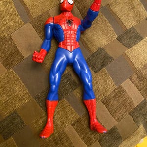 May include: A Spider-Man action figure in a classic red and blue costume. The figure has a red mask, torso, and boots, with blue arms and legs. The figure is posed with one arm raised, displaying a hand gesture.