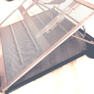 May include: A metal mesh laptop stand with a black base and a silver frame. The stand is angled, designed to elevate a laptop for improved ergonomics and airflow. The mesh design allows for ventilation.
