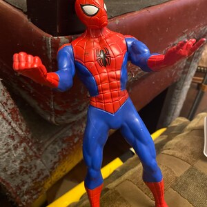 May include: A red, blue, and black Spider-Man action figure. The figure has a red mask with white eyes, a red torso with a black spider emblem, blue arms and legs, and red gloves and boots. The figure is posed with arms outstretched.