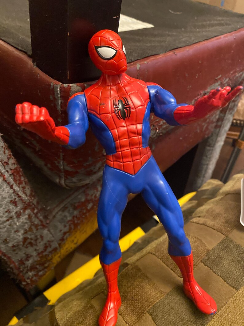 May include: A red, blue, and black Spider-Man action figure. The figure has a red mask with white eyes, a red torso with a black spider emblem, blue arms and legs, and red gloves and boots. The figure is posed with arms outstretched.