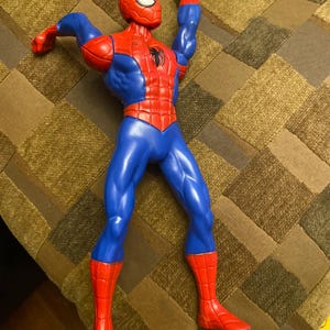 May include: A plastic Spider-Man action figure in a dynamic pose. The figure is primarily red and blue, with a black spider emblem on the chest. The figure's arms are raised, and the head is in a mask.