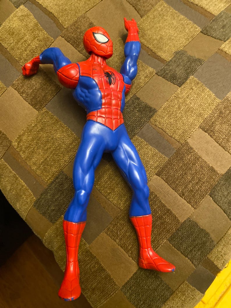 May include: A plastic Spider-Man action figure in a dynamic pose. The figure is primarily red and blue, with a black spider emblem on the chest. The figure's arms are raised, and the head is in a mask.