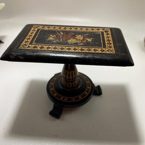 Puede incluir: Una pequeña mesa de madera negra con una tapa cuadrada y un diseño decorativo. La parte superior presenta un patrón incrustado con detalles en marrón y dorado. La mesa tiene una base de pedestal con tres patas pequeñas.