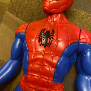 May include: A red and blue Spider-Man action figure. The figure has a red torso with a black spider emblem, blue arms and legs, and a red mask with white eye lenses. The figure is made of plastic.