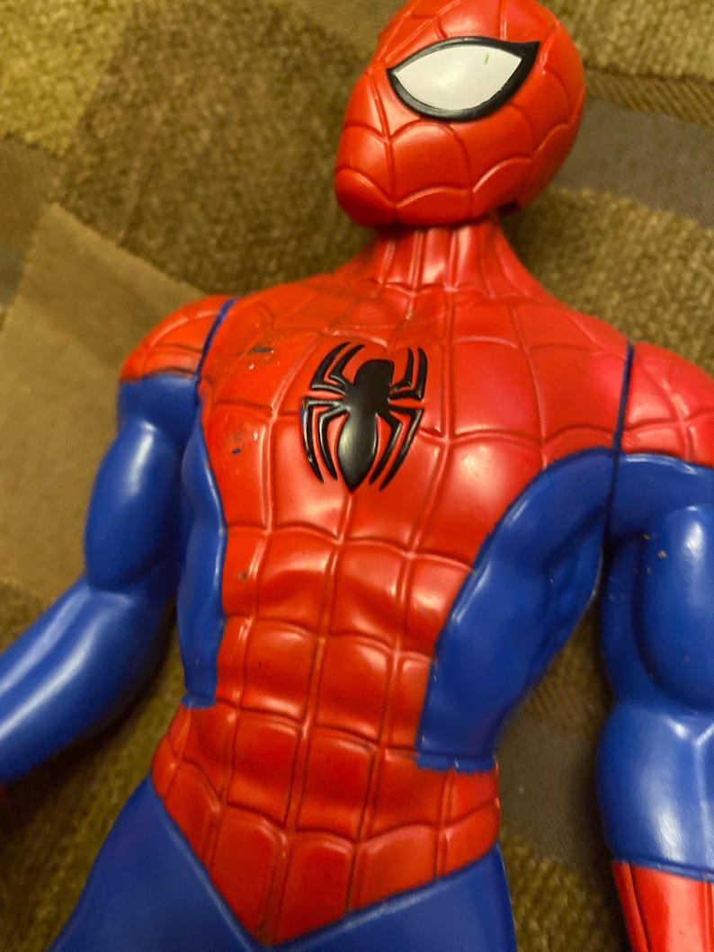 May include: A red and blue Spider-Man action figure. The figure has a red torso with a black spider emblem, blue arms and legs, and a red mask with white eye lenses. The figure is made of plastic.