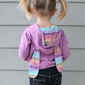 Puede incluir: Una niña pequeña con una sudadera con capucha morada con orejas de conejo a rayas arcoíris en la espalda. Lleva pantalones vaqueros azul oscuro y zapatillas plateadas.