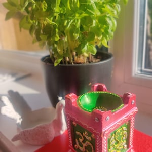 May include: A decorative, square, pink and green candle holder with gold accents and the Om symbol. A white dove figurine and a potted basil plant are also in the image. The candle holder sits on a red surface.