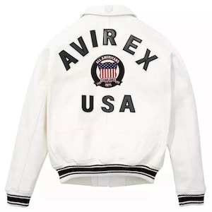 Puede incluir: Chaqueta bomber blanca con pu&ntilde;os y cuello a rayas blancas y negras. La parte trasera presenta la palabra "AVIREX" sobre un emblema circular y la palabra "USA". El emblema tiene un dise&ntilde;o rojo, blanco y azul.