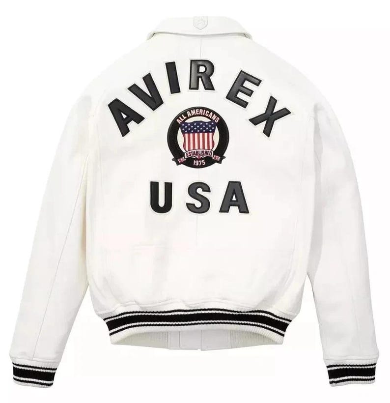 Puede incluir: Chaqueta bomber blanca con pu&ntilde;os y cuello a rayas blancas y negras. La parte trasera presenta la palabra "AVIREX" sobre un emblema circular y la palabra "USA". El emblema tiene un dise&ntilde;o rojo, blanco y azul.