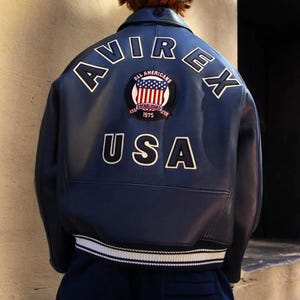 K&ouml;nnte beinhalten: Marineblaue Lederjacke mit den wei&szlig;en Schriftz&uuml;gen "AVIREX" und "USA" auf dem R&uuml;cken. Ein kreisf&ouml;rmiger Aufn&auml;her mit der amerikanischen Flagge und den Worten "ALL AMERICANS ESTABLISHED 1975" ist mittig angebracht. Die Jacke hat einen wei&szlig;en Streifen am unteren Saum.