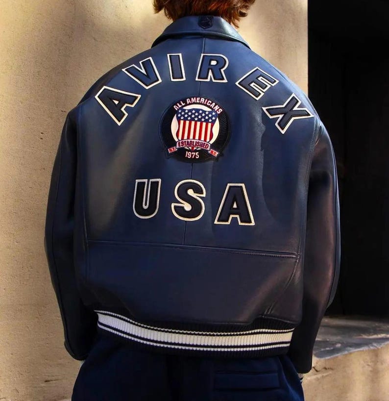 K&ouml;nnte beinhalten: Marineblaue Lederjacke mit den wei&szlig;en Schriftz&uuml;gen "AVIREX" und "USA" auf dem R&uuml;cken. Ein kreisf&ouml;rmiger Aufn&auml;her mit der amerikanischen Flagge und den Worten "ALL AMERICANS ESTABLISHED 1975" ist mittig angebracht. Die Jacke hat einen wei&szlig;en Streifen am unteren Saum.