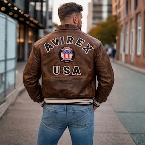 Op de afbeelding: Een bruine leren jas met het woord "AVIREX" over de schouders en "USA" onder een cirkelvormige patch. De jas heeft een geribbelde witte en zwarte gestreepte zoom en manchetten. De persoon draagt een blauwe spijkerbroek.