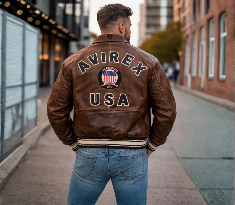 Op de afbeelding: Een bruine leren jas met het woord "AVIREX" over de schouders en "USA" onder een cirkelvormige patch. De jas heeft een geribbelde witte en zwarte gestreepte zoom en manchetten. De persoon draagt een blauwe spijkerbroek.